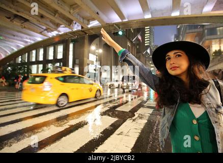 Schöne, modische junge Frau, die in einer regnerischen, stürmischen Nacht in Manhattan, New York City, NY, ein Taxi in der 42. Street vor der Grand Central Station anhagelt Stockfoto