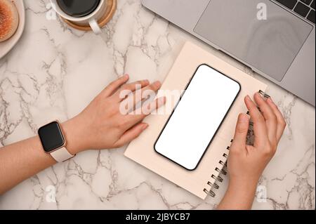 Die Frau, die ihr Smartphone mit weißem Bildschirm auf einem Spiralnotepad auf einem modernen Marmortisch ablegt. Stockfoto