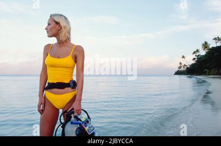 Junge Frau, die am tropischen Strand steht und ihre Tauchausrüstung hält Stockfoto