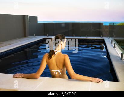Rückansicht einer jungen Frau, die im Swimmingpool stand und sich gegen den Rand lehnte, mit Blick auf das Meer über die Wand Stockfoto