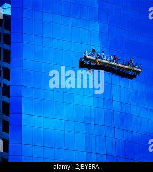 Zwei Männer in hängenden Gerüsten, die die Fenster eines Hochhauses aus Glas putzen und Wolken in Glas spiegeln Stockfoto