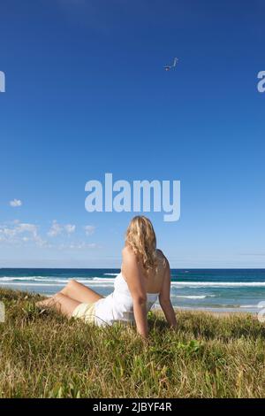 Rückansicht einer jungen Frau, die am Strand sitzt und Drachen fliegt Stockfoto