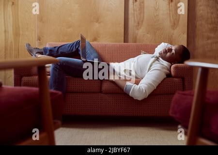 Junger Mann, der mit seinem Computer auf dem Schoß ein Nickerchen auf dem Loveseat macht Stockfoto