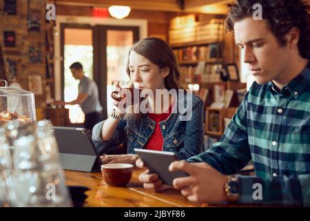 Junger Mann und Frau, die an der Theke im Café-Buchladen mit digitalen Tabletten sitzen, Frau, die einen Schluck Kaffee trinkt Stockfoto