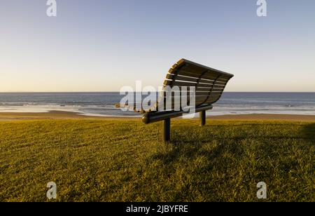 Leere Parkbank auf einer grasbewachsenen Bank mit Blick auf den Strand am frühen Morgen Stockfoto
