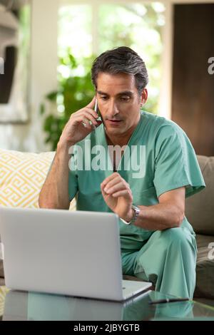 Hispanischer Arzt, der von zu Hause aus Telemedizin praktiziert, Handy und Laptop benutzt und mit dem Patienten während eines Videoanrufs spricht Stockfoto