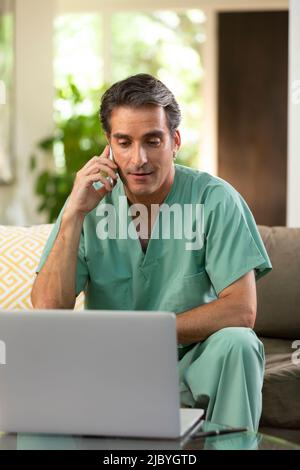 Hispanischer Arzt, der von zu Hause aus Telemedizin praktiziert, Handy und Laptop benutzt und mit dem Patienten während eines Videoanrufs spricht Stockfoto