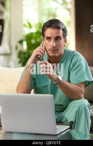 Hispanischer Arzt, der von zu Hause aus Telemedizin praktiziert, Handy und Laptop benutzt und dem Patienten bei einem Videoanruf zugehört Stockfoto