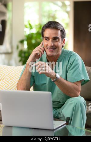 Hispanischer Arzt, der von zu Hause aus Telemedizin praktiziert, Handy und Laptop benutzt und dem Patienten bei einem Videoanruf zugehört Stockfoto