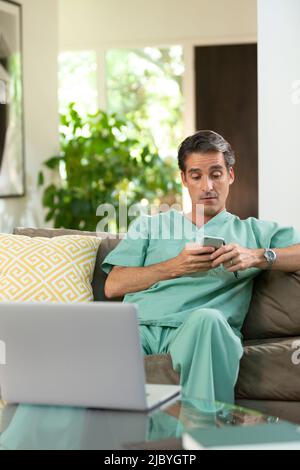 Hispanischer Arzt, der von zu Hause aus Telemedizin praktiziert, Handy und Laptop benutzt und dem Patienten bei einem Videoanruf zugehört Stockfoto