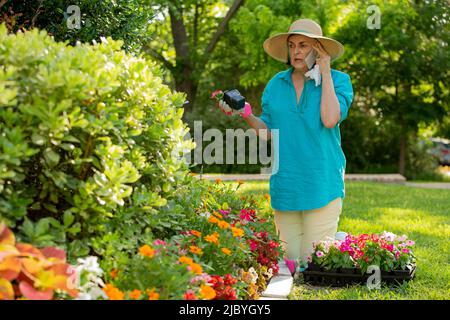 Ältere kaukasische Frau, die in ihrem Garten arbeitet, Blumen vor dem Haus pflanzt und ihren Arzt wegen ihrer saisonalen Allergien anruft Stockfoto