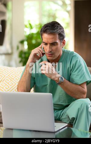 Hispanischer Arzt, der von zu Hause aus Telemedizin praktiziert, Handy und Laptop benutzt und dem Patienten bei einem Videoanruf zugehört Stockfoto