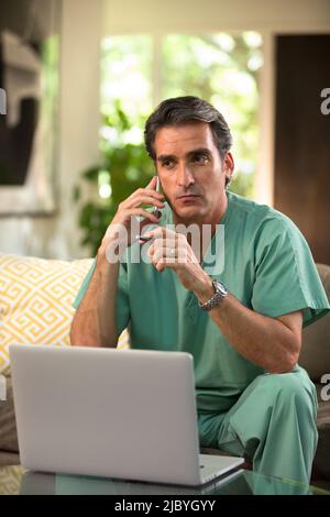 Hispanischer Arzt, der von zu Hause aus Telemedizin praktiziert, Handy und Laptop benutzt und dem Patienten bei einem Videoanruf zugehört Stockfoto