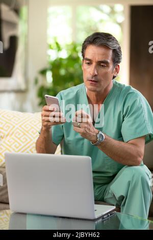 Hispanischer Arzt, der von zu Hause aus Telemedizin praktiziert, Handy und Laptop benutzt und dem Patienten bei einem Videoanruf zugehört Stockfoto