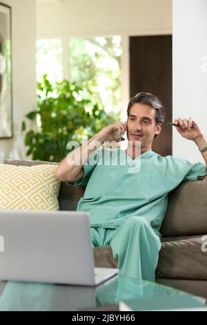 Hispanischer Arzt, der von zu Hause aus Telemedizin praktiziert, Handy und Laptop benutzt und dem Patienten bei einem Videoanruf zugehört Stockfoto