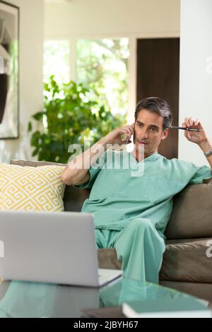Hispanischer Arzt, der von zu Hause aus Telemedizin praktiziert, Handy und Laptop benutzt und dem Patienten bei einem Videoanruf zugehört Stockfoto