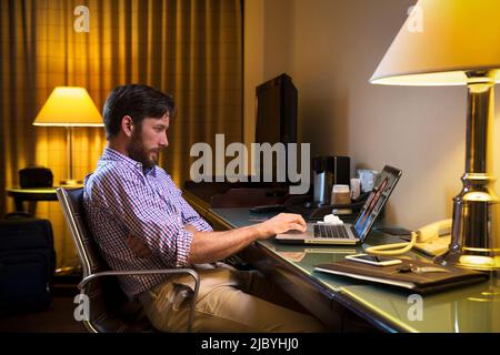 Kaukasischer Mann, der spät in einem Hotelzimmer arbeitet und sich krank fühlt, so dass er sich mit einem Arzt auf einem Laptop telemedizinisch trifft Stockfoto