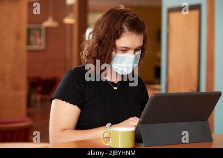 Der jungen kaukasischen Frau trägt schwarzes T-Shirt und Gesichtsmaske, sitzt an der Bar in der Küche der Innenstadt Loft mit Kaffeebecher mit Tablet mit Tastatur Stockfoto
