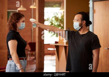 Die junge kaukasische Frau trägt ein schwarzes T-Shirt und eine Gesichtsmaske und lässt die Temperatur mit einem berührungslosen Thermometer von einem maskierten Mitarbeiter überprüfen Stockfoto