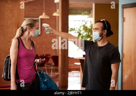 Kaukasische Frau mittleren Alters mit rosa Tank-Top- und Gesichtsmaske, die die Temperatur mit einem berührungslosen Thermometer von einem maskierten Mitarbeiter überprüft hat Stockfoto