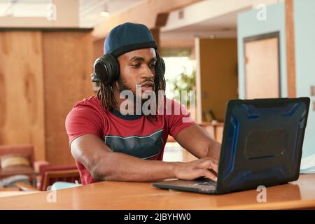 Porträt eines jungen ethnischen Mannes mit rotem T-Shirt und Strickmütze mit Kopfhörern, der an der Bar in der Küche des Loft in der Innenstadt mit einem Laptop-Computer sitzt Stockfoto