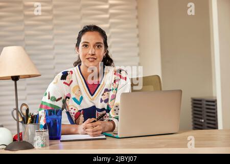 Porträt einer jungen ethnischen Frau, die mit einem Laptop-Computer am Schreibtisch sitzt und Notizen auf einem Notizblock schreibt Stockfoto
