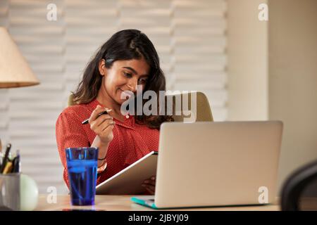 Porträt einer jungen ethnischen Frau, die mit einem Laptop-Computer am Schreibtisch sitzt und Notizen auf einem Notizblock schreibt Stockfoto