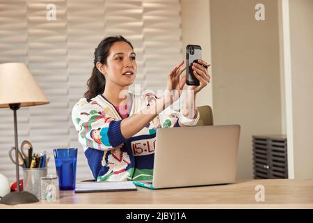 Junge ethnische Frau, die am Schreibtisch sitzt, mit einem Laptop-Computer, der Notizen auf einem Notizblock gemacht hat und ein Smartphone benutzt Stockfoto
