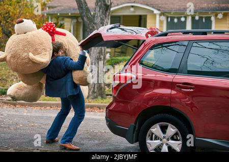 Ein kitschiger junger Mann, der einen überdimensionalen Teddybären herausziehen muss und über sich stolpert. Stockfoto