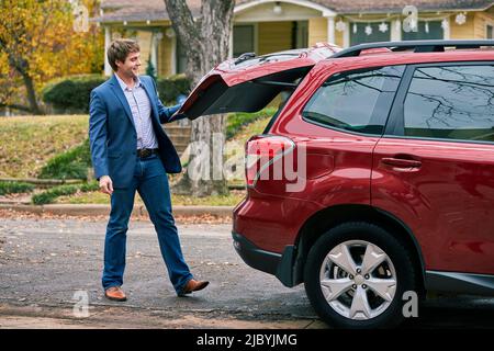 Weitwinkelaufnahme eines Mannes, der zum Kofferraum seines Autos läuft und seinen Partner mit einem übergroßen Teddybären überrascht. Stockfoto