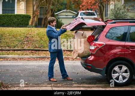 Ein kooffiger junger Mann, der auf den Rücken seines roten Autos geht, den Kofferraum öffnet und einen überdimensionalen Teddybären anschaut, der seinen Kopf aus dem Lastwagen herausknallt. Stockfoto