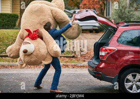 Breiter Schuss eines Mannes, der einen überdimensionalen Teddybären über seine Schulter hält und den Kofferraum seines Autos schließt. Stockfoto