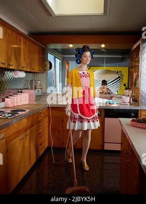 Frau saugt die Küche, während sie Haarwalzen trägt, in einer Küche im Stil der Mitte des Jahrhunderts. Stockfoto
