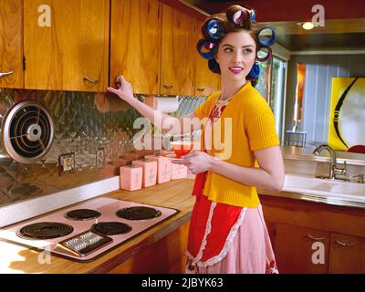 Porträt einer Frau, die in einer Küche mit Lockenwicklern in den Haaren in einer Küche im Stil der Mitte des Jahrhunderts einen Schrank öffnet. Stockfoto