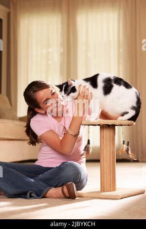 Teenager kaukasisches Mädchen, das im Wohnzimmer mit gekreuzten Beinen auf dem Teppich sitzt und ihre Katze streichelt, die Katze sitzt auf einem Kratzbaum Stockfoto