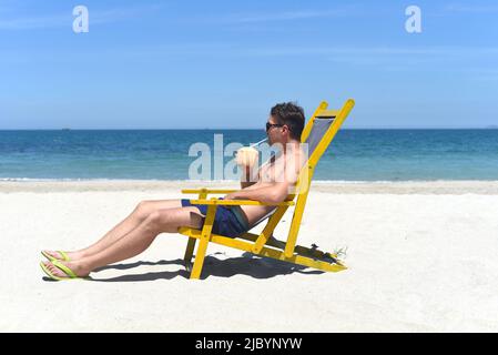 Ein junger glücklicher Mann trinkt Kokosnusssaft, der auf einem Liegestuhl an einem südchinesischen Strand in vietnam sitzt Stockfoto