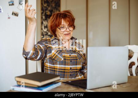 Ältere überraschte Frau, die eine Brille trug, am Tisch saß, die Hand in der Nähe von Laptop-Büchern zu Hause hob, gestikulte, ausrief. Stockfoto