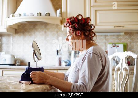 Seitenansicht einer älteren Frau, die pinkfarbene Haarlockenwickler aus Kunststoff aufsetzt und am Tisch in der beigen Küche sitzt und auf den Spiegel schaut. Stockfoto