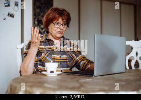 Ältere, staunende Frau, die kariertes Hemd und Brille trägt, am Tisch auf einem Stuhl sitzt, die Hand hebt und auf den Laptop schaut. Stockfoto