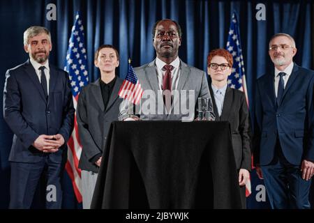 Porträt eines reifen afrikanischen Führers, der bei der Pressekonferenz auf der Tribüne mit seinem Geschäftsteam hinter ihm stand Stockfoto