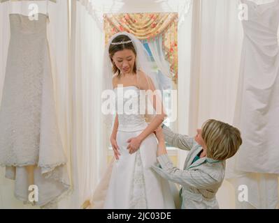 Junge Braut, die versuchen, auf ihr Kleid mit Hilfe eines Mitarbeiters Stockfoto