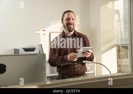 Der junge Mann blickt aus dem Fenster seines Büros und hält ein Tablet. Stockfoto