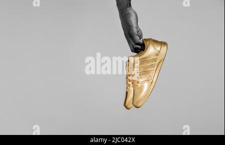 Entsättigte/detailgezeichnete Aufnahme der Hand eines schwarzen Mannes mit goldlackierten Sneakers vor dem weißen Brett. Stockfoto