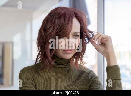 Eine Frau mittleren Alters mit roten Haaren schaut aus dem Fenster und dreht ihre Haare. Stockfoto