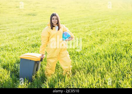 Eine Frau in einem gelben Schutzanzug steht mitten auf einem grünen Feld und hält einen Globus und trägt mit ihrer anderen Hand eine Mülltonne. Stockfoto