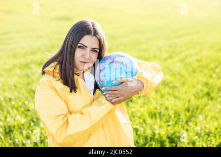 Eine Frau in einem gelben Schutzanzug steht in der Mitte eines grünen Feldes und hält einen Globus. Stockfoto