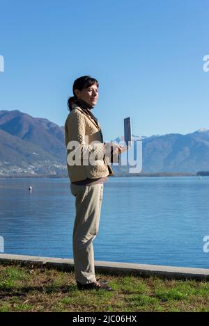 Elegante Frau mit Laptop auf der Waterfront mit Berg in Ascona, Schweiz. Stockfoto