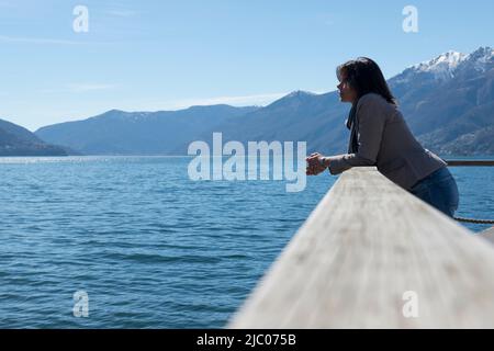 Frau steht und schief auf einem Geländer an der Waterfront zu einem alpinen Lago Maggiore mit Berg in Ascona, Schweiz. Stockfoto