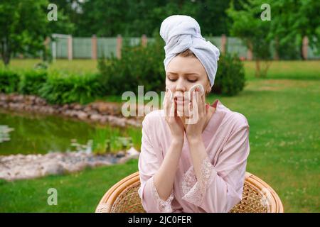 Schöne Frau mit perfekter Haut Make-up und Reinigung der Haut auf ihrem Gesicht mit Baumwollkissen nach der Dusche in Handtuch auf dem Kopf, während im Stuhlgarten zu Hause in Frischluft Hautpflege-Konzept sitzen Stockfoto