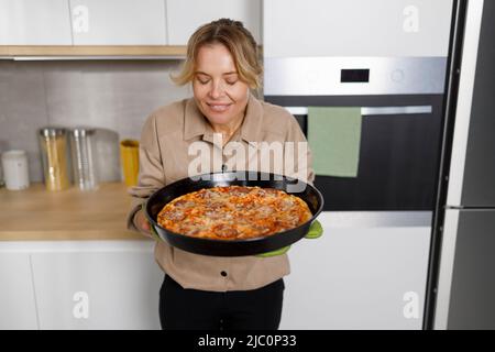 Frau mittleren Alters hält und schnüffelt ein Backblech mit frisch gebackener Pizza Stockfoto